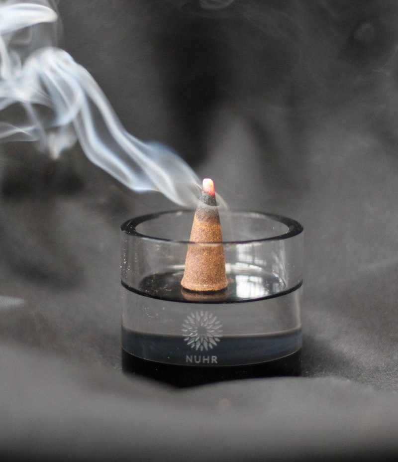 A lit NUHR oud incense cone releasing a curl of smoke in a glass holder